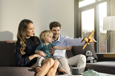 Glückliche Eltern und ihr Sohn spielen mit einem Holzspielzeugflugzeug auf dem Sofa zu Hause - SBOF01273