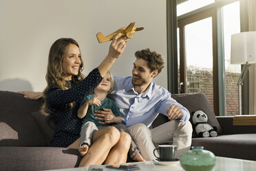 Glückliche Eltern und ihr Sohn spielen mit einem Holzspielzeugflugzeug auf dem Sofa zu Hause - SBOF01274