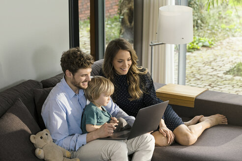 Lächelnde Eltern und ihr Sohn sitzen auf dem Sofa und benutzen einen Laptop zu Hause - SBOF01280