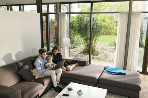 Parents and son sitting on sofa in modern living room using laptop at home - SBOF01281