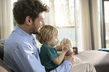 Father and son playing with teddy bear on couch at home - SBOF01282