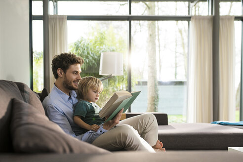Father and son reading book on couch at home - SBOF01284