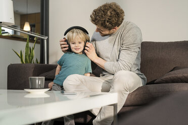 Father putting on headphones on son on couch at home - SBOF01290