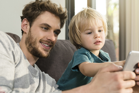 Vater und Sohn schauen zu Hause auf der Couch auf ihr Smartphone, lizenzfreies Stockfoto