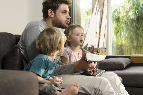 Vater und Kinder blasen in die Segel eines Spielzeugmodellschiffs auf der Couch zu Hause - SBOF01297
