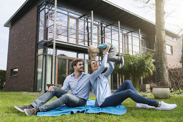 Happy parents with son sitting on blanket in garden in front of their home - SBOF01315