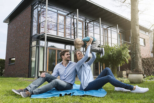 Glückliche Eltern mit Sohn sitzen auf einer Decke im Garten vor ihrem Haus - SBOF01315