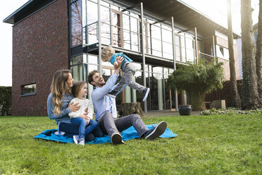 Happy family on blanket in garden in front of their home with father lifting up son - SBOF01327