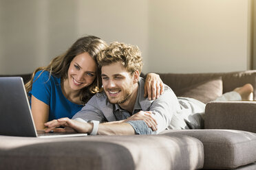 Smiling couple using laptop lying on sofa at home - SBOF01331