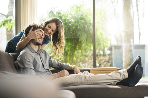 Smiling woman covering her boyfriend's eyes on sofa at home stock photo