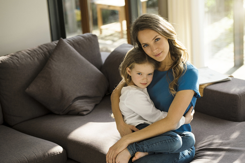 Porträt einer lächelnden Mutter, die ihre Tochter auf dem Sofa zu Hause im Arm hält, lizenzfreies Stockfoto