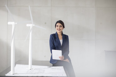 Portrait of smiling young woman with blueprint, wind turbine models and tablet in office - ZEF14918