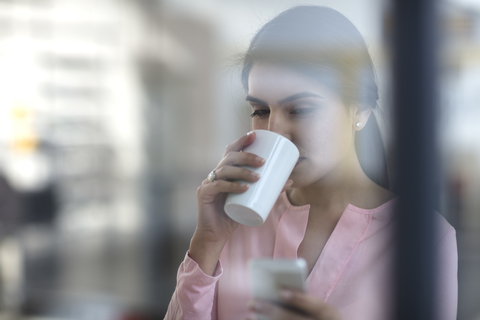 Porträt einer jungen Frau mit Mobiltelefon und Kaffee im Büro, lizenzfreies Stockfoto