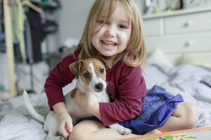 Porträt eines glücklichen kleinen Mädchens, das mit einem Jack Russel Terrier-Welpen auf dem Bett hockt - KMKF00142