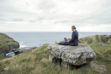 UK, Cornwall, Tintagel, Geschäftsmann sitzt auf einem Felsen an der Küste und benutzt einen Laptop - PSTF00084
