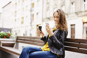 Porträt einer glücklichen Frau, die mit einer Eistüte auf einer Bank sitzt und auf ihr Mobiltelefon schaut - BSZF00205