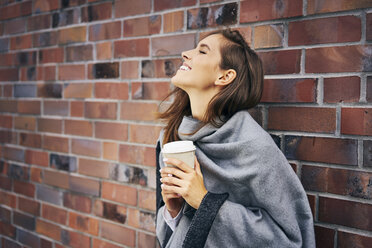 Glückliche Frau mit Kaffee zum Mitnehmen vor einer Backsteinmauer - BSZF00207