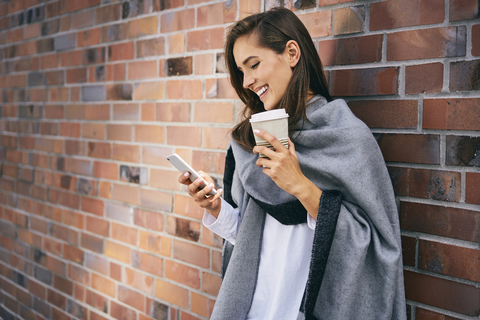 Happy woman with coffee to go looking at cell phone in front of brick wall stock photo