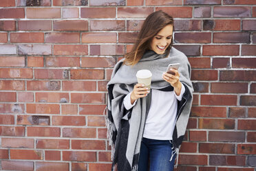 Glückliche Frau mit Kaffee zum Mitnehmen, die vor einer Backsteinmauer auf ihr Mobiltelefon schaut - BSZF00210