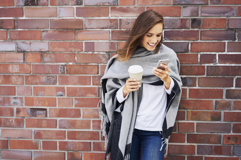 Happy woman with coffee to go looking at cell phone in front of brick wall - BSZF00210