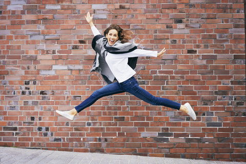 Glückliche junge Frau, die vor einer Backsteinmauer in die Luft springt, lizenzfreies Stockfoto