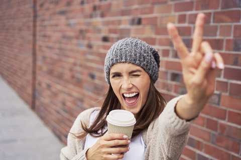 Porträt einer jungen Frau mit Kaffee zum Mitnehmen und Siegeszeichen, lizenzfreies Stockfoto