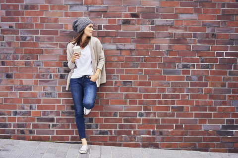 Junge Frau mit Kaffee zum Mitnehmen lehnt an einer Backsteinmauer, lizenzfreies Stockfoto