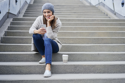 Happy woman with coffee to go sitting on stairs looking at cell phone stock photo