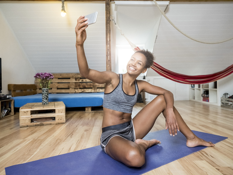 Lächelnde junge Frau, die auf einer Yogamatte sitzt und ein Selfie macht, lizenzfreies Stockfoto