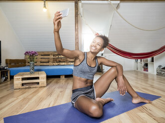 Lächelnde junge Frau, die auf einer Yogamatte sitzt und ein Selfie macht - MADF01387