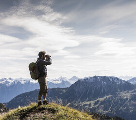 Österreich, Tirol, junger Mann steht in einer Berglandschaft und betrachtet die Aussicht mit einem Fernglas - UUF12574