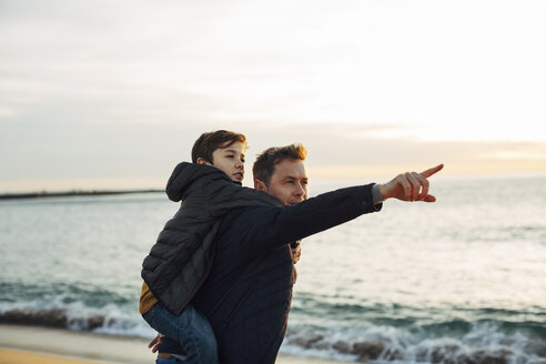 Vater trägt Sohn huckepack am Strand bei Sonnenuntergang und zeigt mit dem Finger - EBSF02024