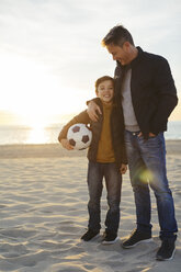 Father embracing son with football on the beach at sunset - EBSF02037