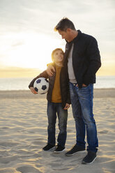 Father embracing son with football on the beach at sunset - EBSF02038