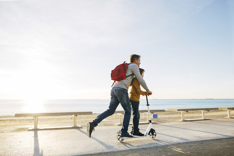 Vater und Sohn fahren Roller auf der Strandpromenade bei Sonnenuntergang, lizenzfreies Stockfoto