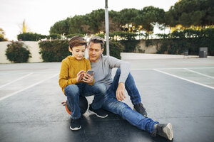 Vater und Sohn sitzen auf einem Basketballplatz im Freien und benutzen ein Mobiltelefon - EBSF02068