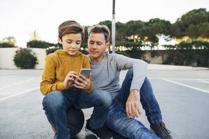 Vater und Sohn sitzen auf einem Basketballplatz im Freien und benutzen ein Mobiltelefon - EBSF02069