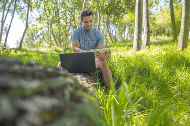 Mann mit Laptop sitzt auf einem Baumstamm in der Natur und benutzt ein Tablet - ZEF14942