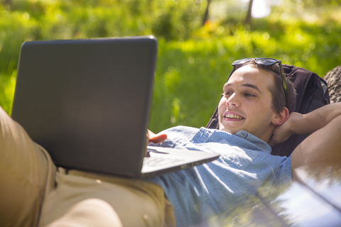 Porträt eines glücklichen jungen Mannes, der in der Natur auf einem Baumstamm liegt und einen Laptop benutzt, lizenzfreies Stockfoto
