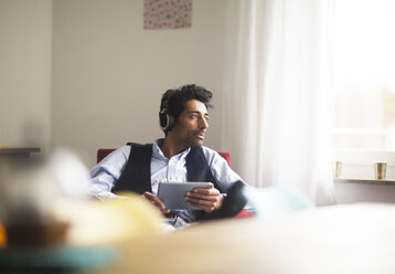 Portrait of pensive man with headphones and tablet sitting on armchair looking out of window - SGF02148