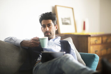 Portrait of pensive man with coffee mug and tablet sitting on couch at home - SGF02150