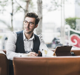 Junger Mann arbeitet in einem Café mit Tablet und macht sich Notizen - UUF12598