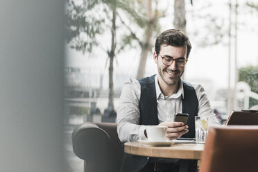 Lächelnder junger Mann, der in einem Café arbeitet und auf sein Handy schaut - UUF12601