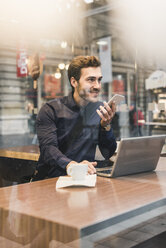 Smiling young businessman in a cafe at train station with cell phone and laptop - UUF12639