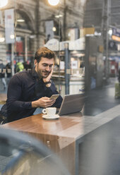 Young businessman in a cafe at train station with cell phone and laptop - UUF12641