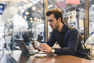 Young businessman in a cafe at train station with cell phone and laptop - UUF12644