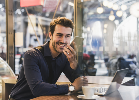 Lächelnder junger Geschäftsmann in einem Café am Bahnhof mit Handy und Laptop, lizenzfreies Stockfoto