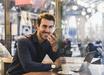 Lächelnder junger Geschäftsmann in einem Café am Bahnhof mit Handy und Laptop - UUF12645