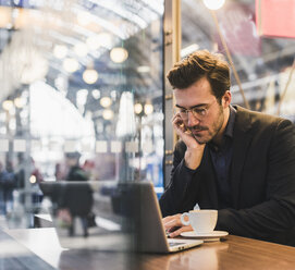 Junger Geschäftsmann in einem Café am Bahnhof mit einer Tasse Kaffee und einem Laptop - UUF12646