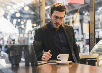 Young businessman in a cafe at train station with cup of coffee, laptop and cell phone - UUF12647